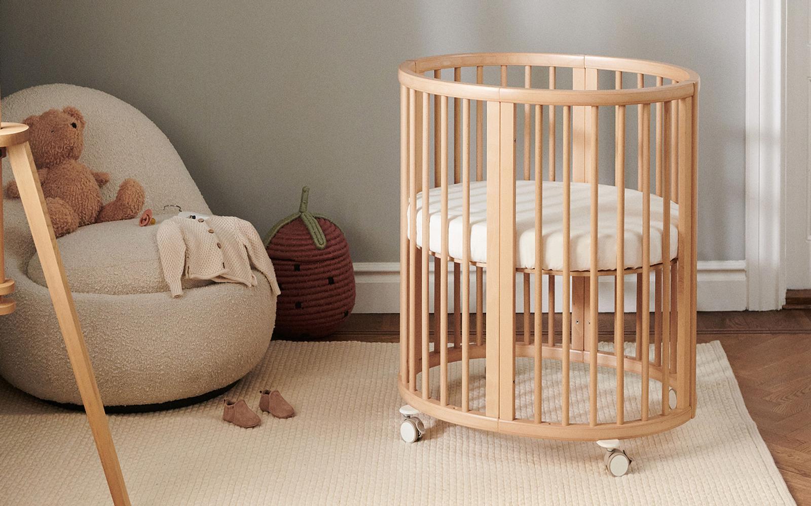 A round wooden crib on wheels sits on a rug in a nursery. An armchair holds a teddy bear and baby clothes.