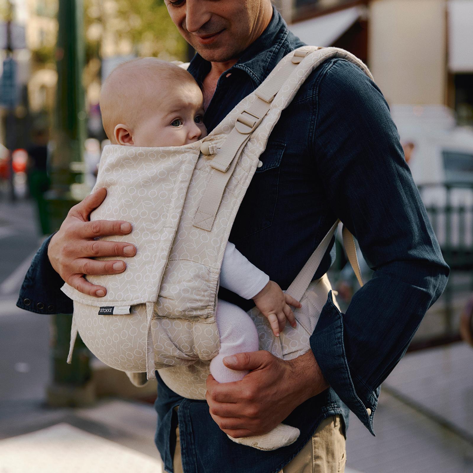 A man smiles, carrying a baby in a beige patterned STOKKE baby carrier outdoors.