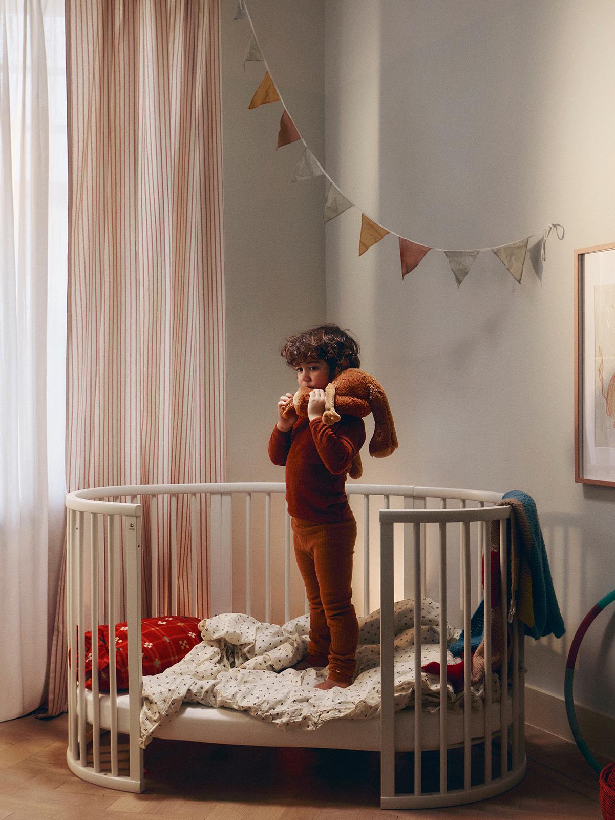 錆色のパジャマを着た幼児がStokke Sleepiベビーベッドの中に立ち、茶色のぬいぐるみウサギを抱きしめている。