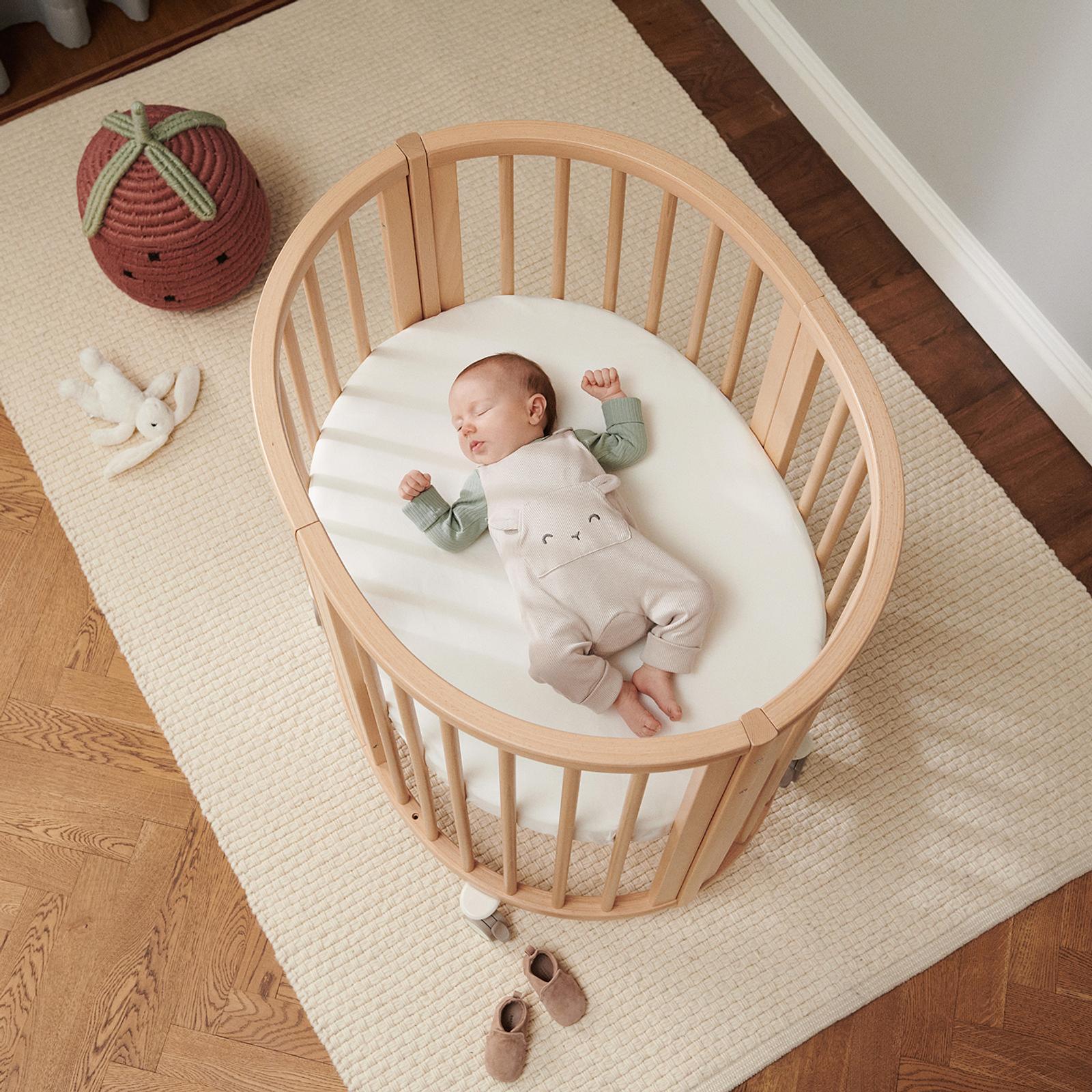 Ein friedliches Baby schläft in einem ovalen Holzbettchen, mit einem Hasenspielzeug und einem Erdbeerkissen in der Nähe.