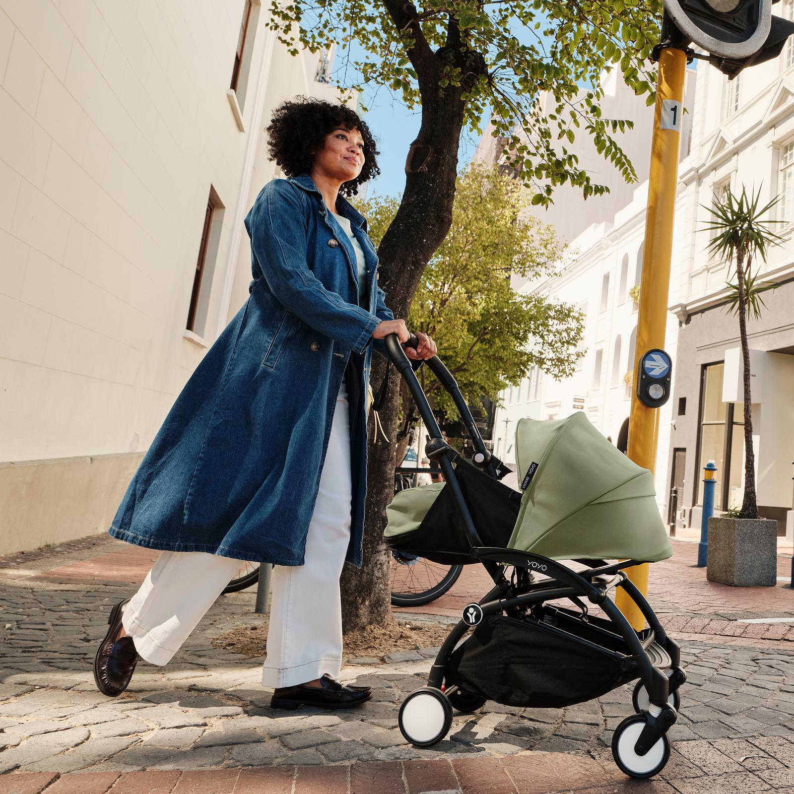 笑顔の女性が晴れた街の通りで緑色のSTOKKE YOYOベビーカーを押しています。