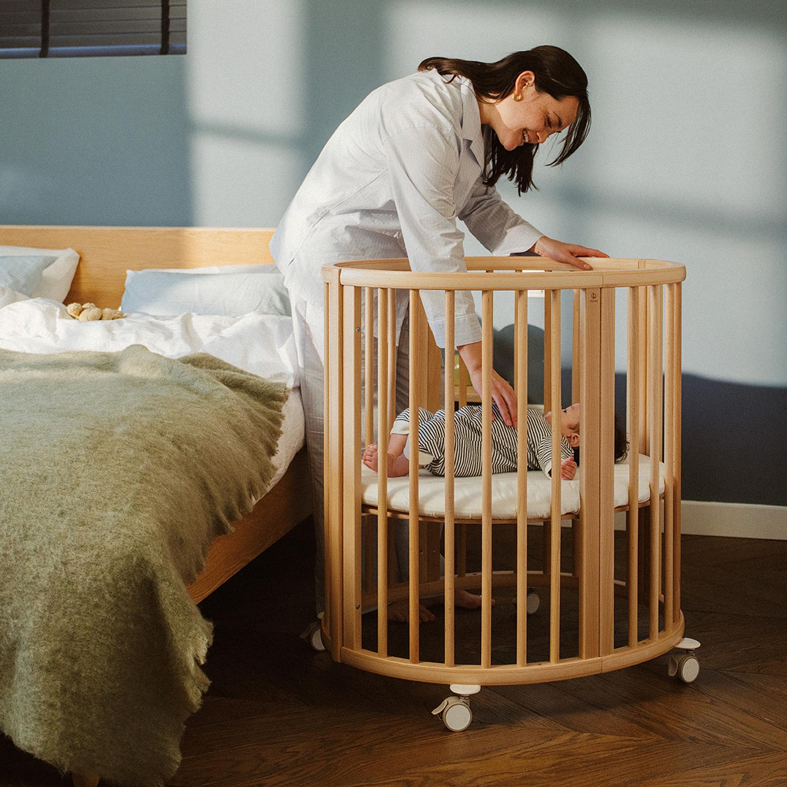 A smiling woman in pajamas gently touches a baby sleeping in a round STOKKE Sleepi Mini crib.