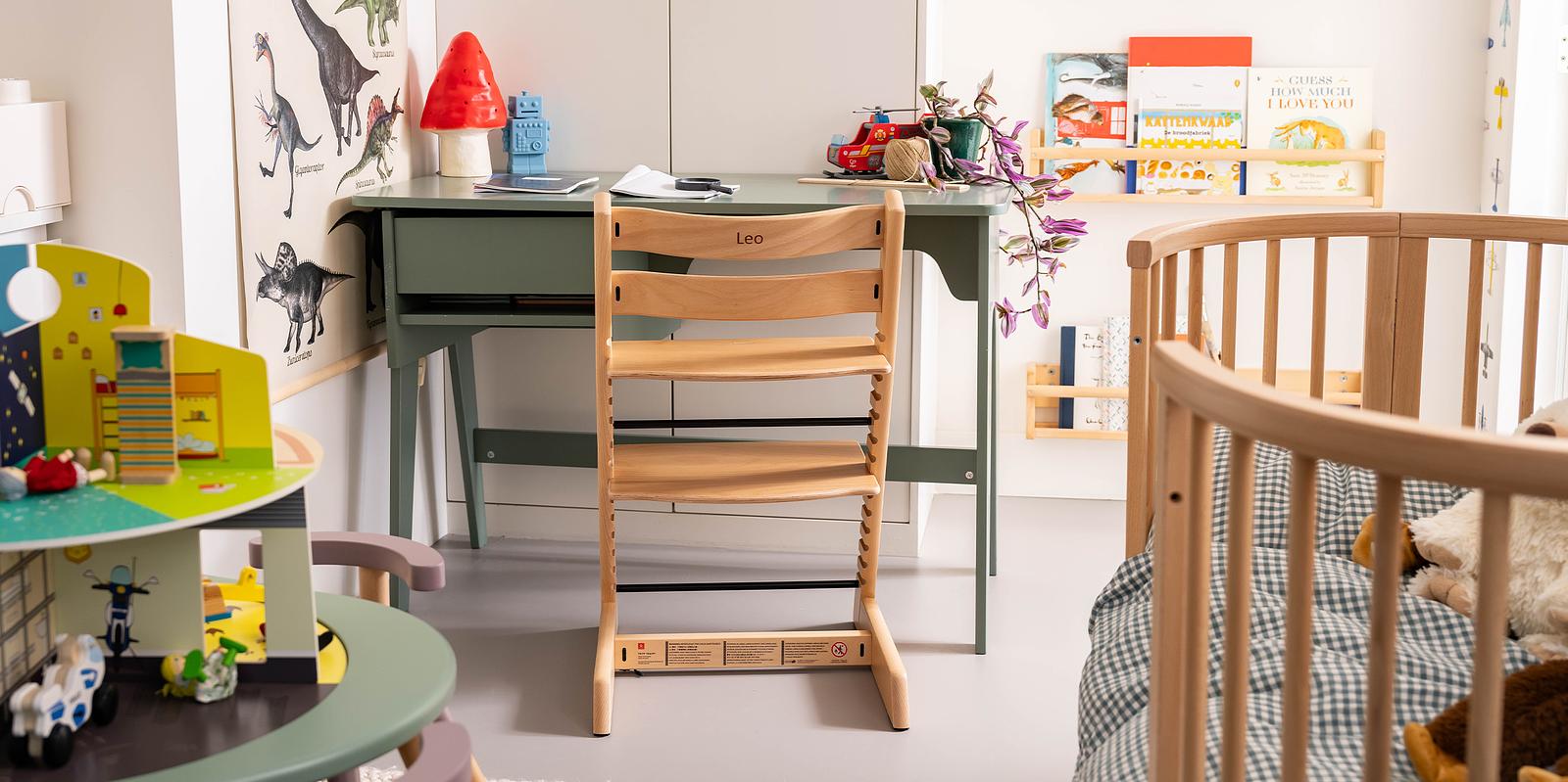 A natural wood Tripp Trapp chair with 'Leo' at a green desk in a child's room with a crib.