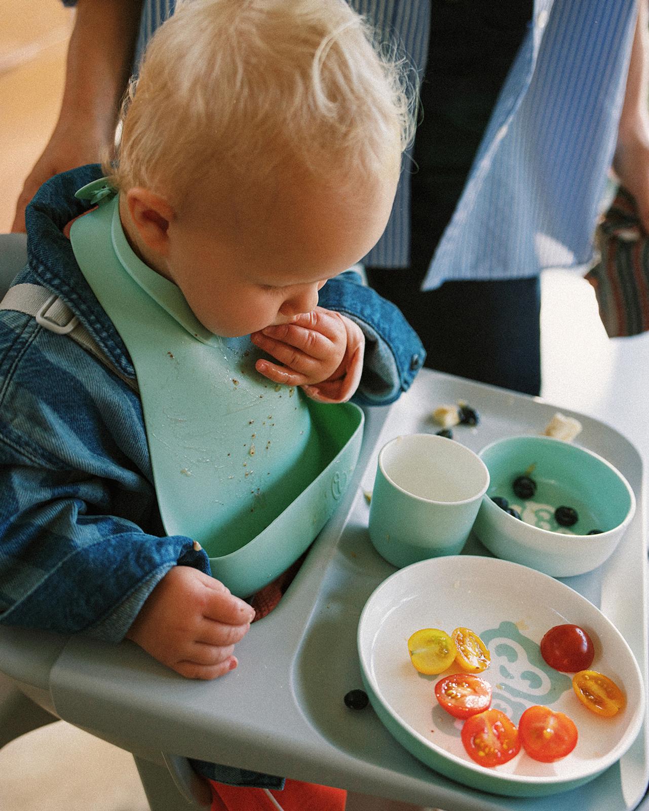 Bambino biondo in un seggiolone grigio, indossa una giacca di jeans e un bavaglino color menta, mangia pomodorini e mirtilli.