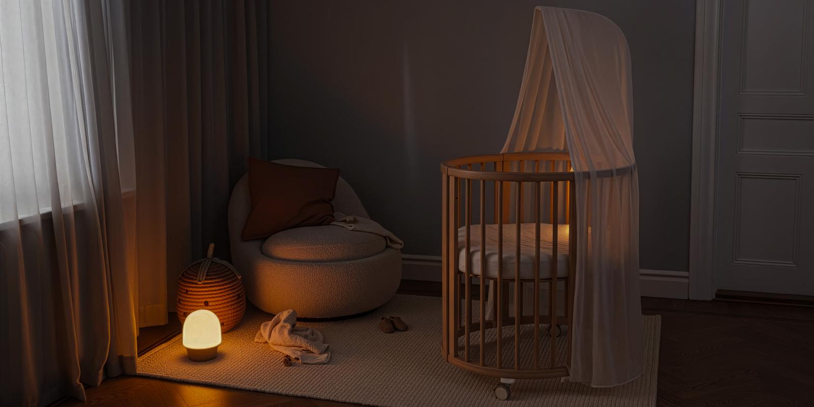 薄暗いナーサリールーム。STOKKE Sleepiベビーベッドには天蓋があり、光る常夜灯とアームチェアが置かれている。