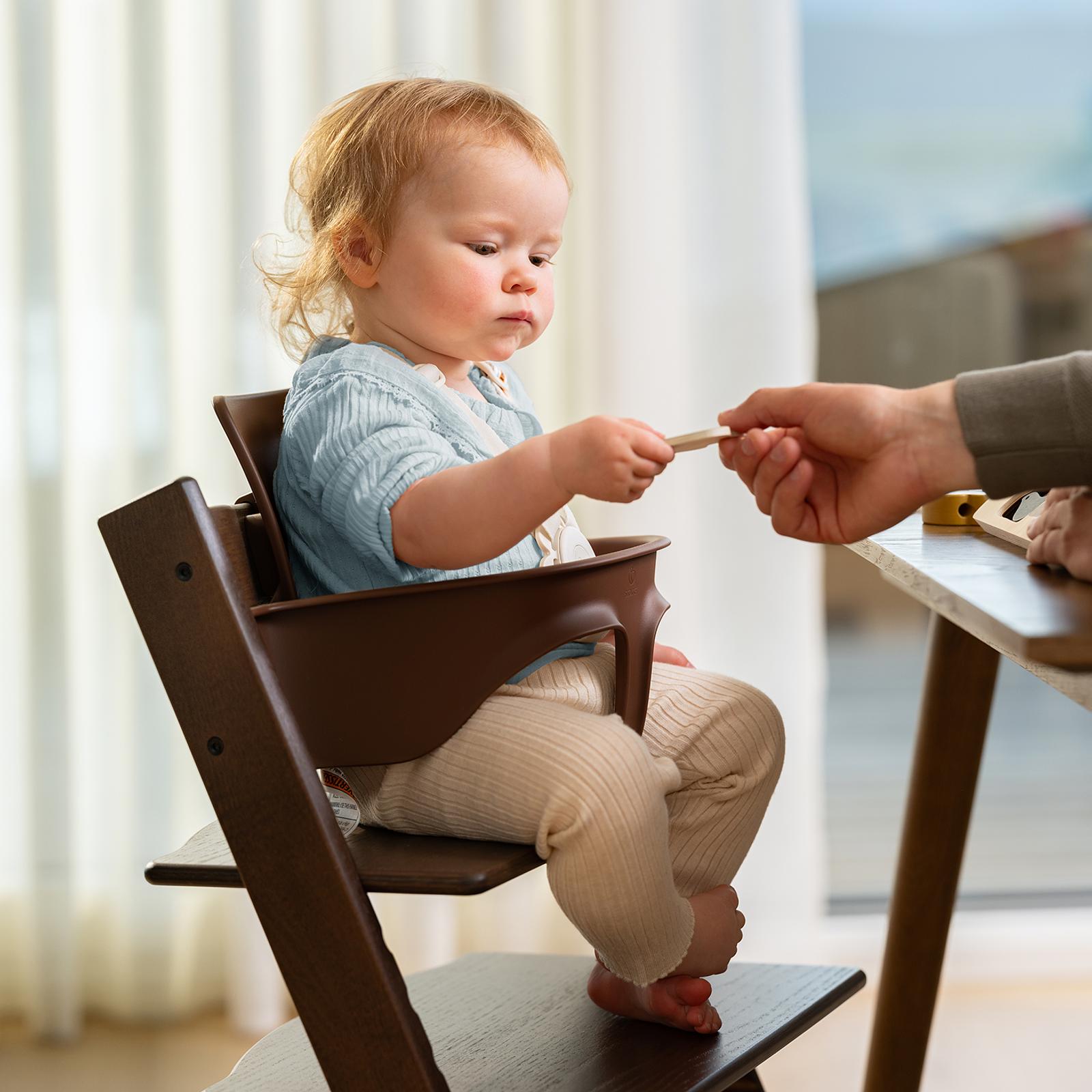 Toddler in a brown Tripp Trapp high chair takes a wooden toy from an adult's hand.