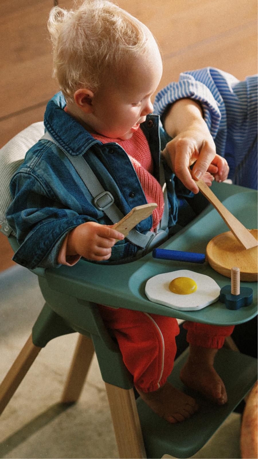 Un bambino biondo con una giacca di jeans gioca con cibo giocattolo di legno in un seggiolone verde, aiutato dalla mano di un adulto.