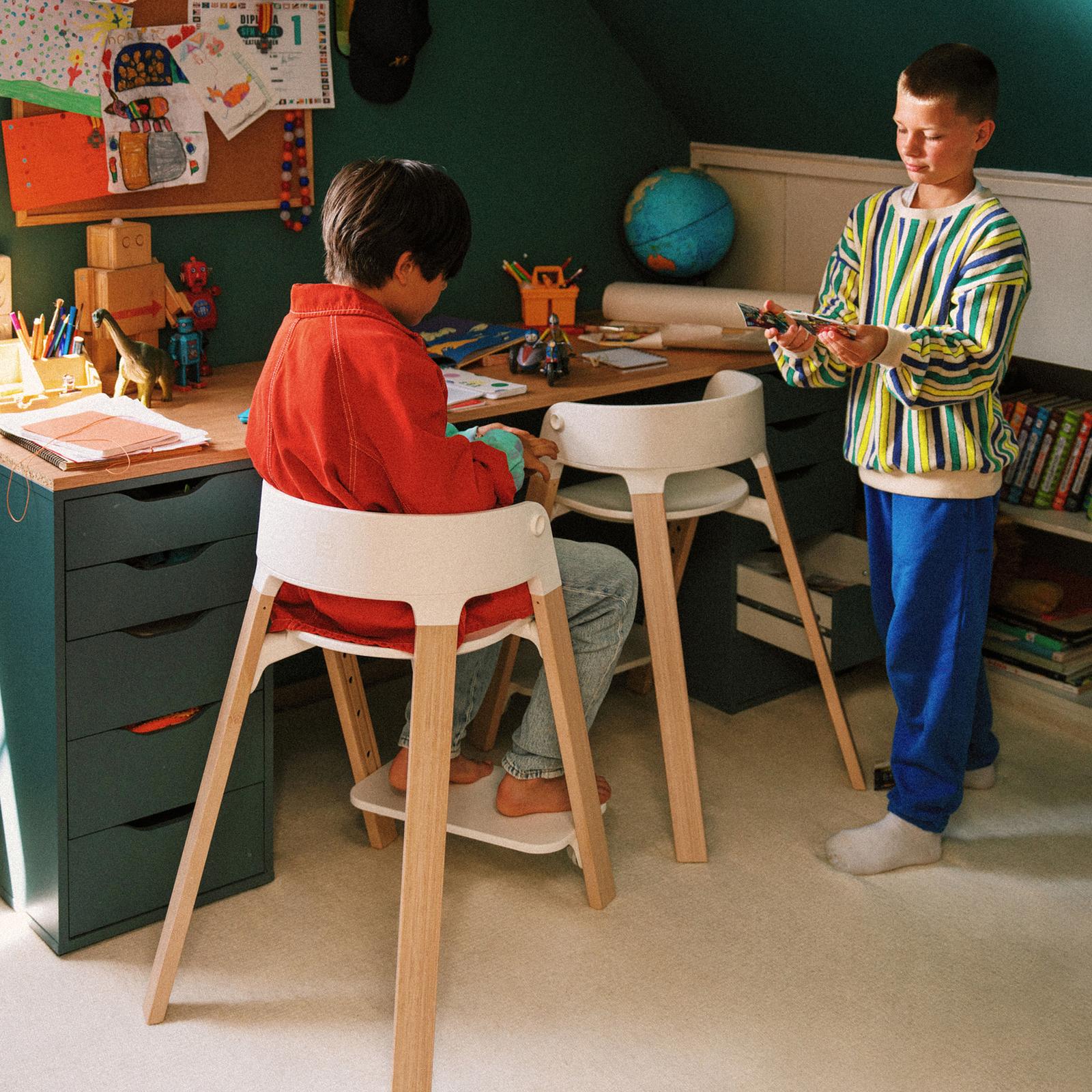 Two boys in a playroom. One draws at a desk in a child's chair, the other stands holding cards.