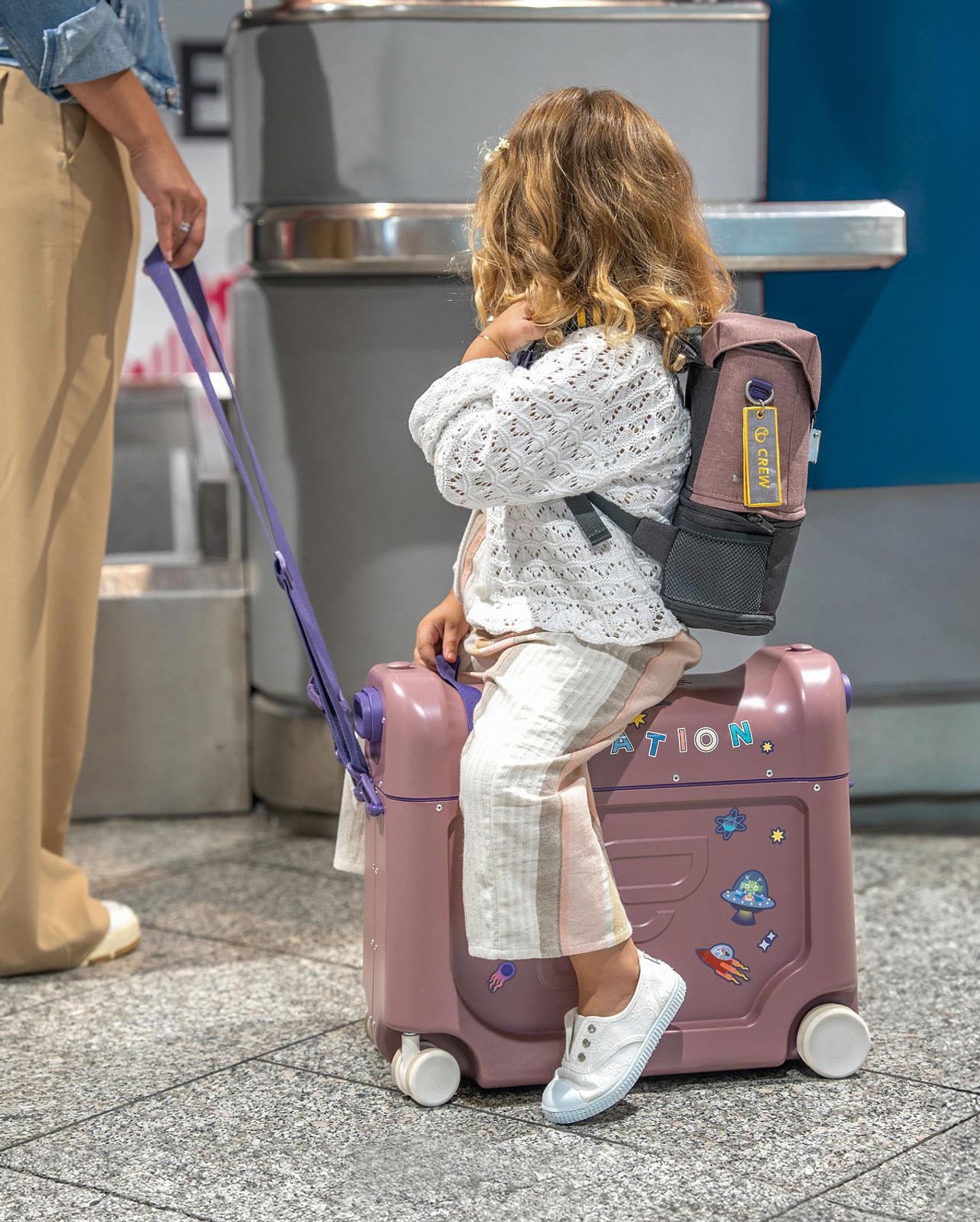 A child sits on a purple ride-on suitcase with stickers, pulled by an adult in an airport.