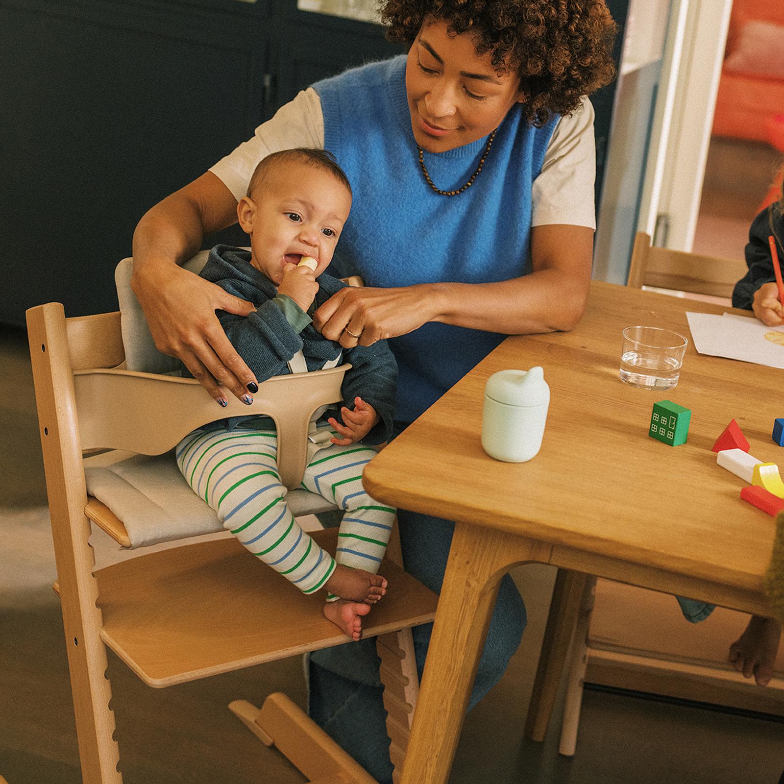 Woman helps baby eating in a Tripp Trapp high chair at a wooden table with toys.