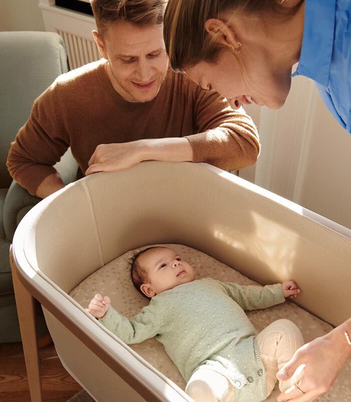 Mother and father looking at their newborn baby in snoozi bassinet color sandy beige.