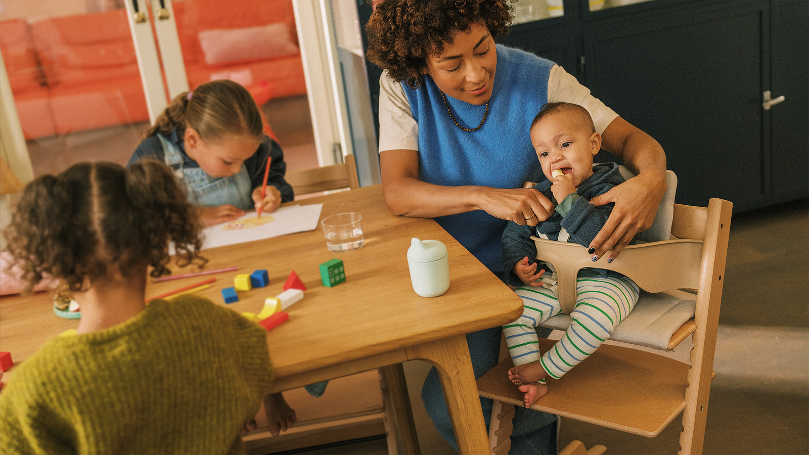 A mother helps a baby in a Tripp Trapp high chair while two older children play and draw.