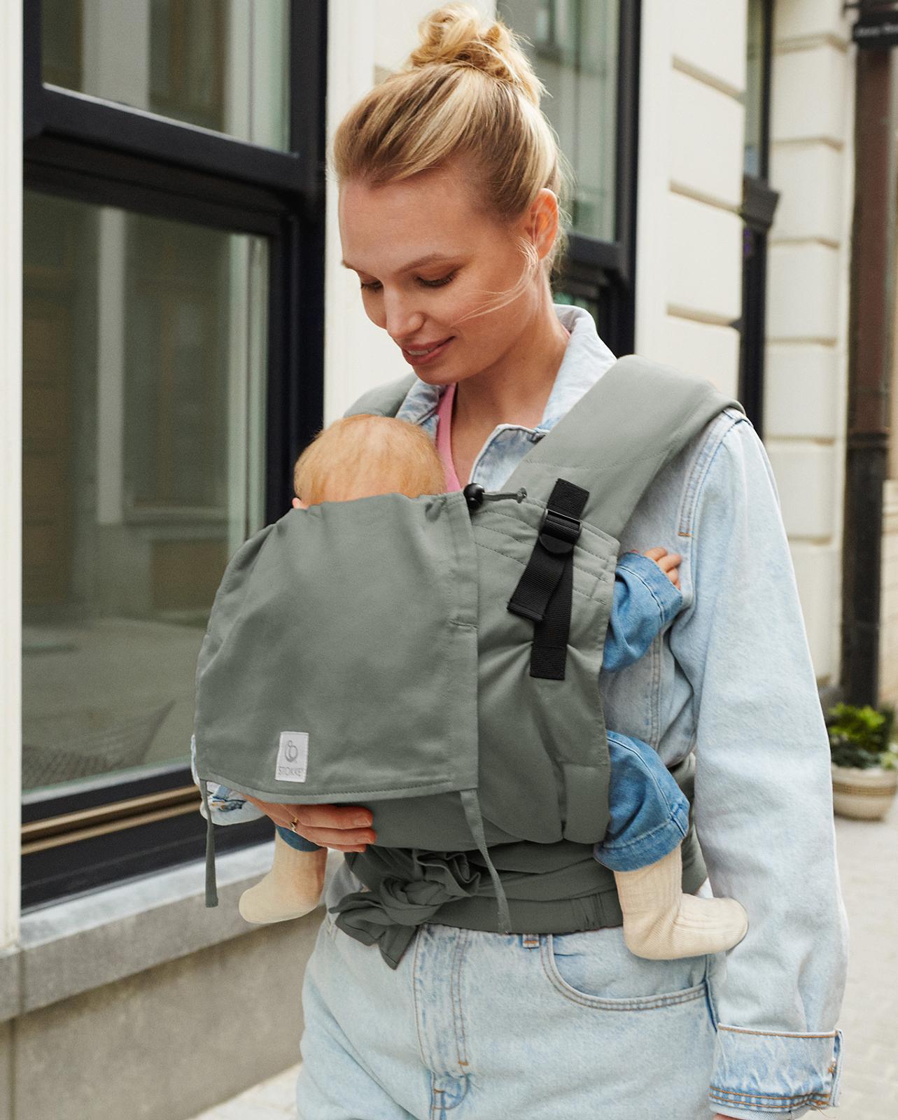 A smiling woman carries a baby in a grey baby carrier outdoors.