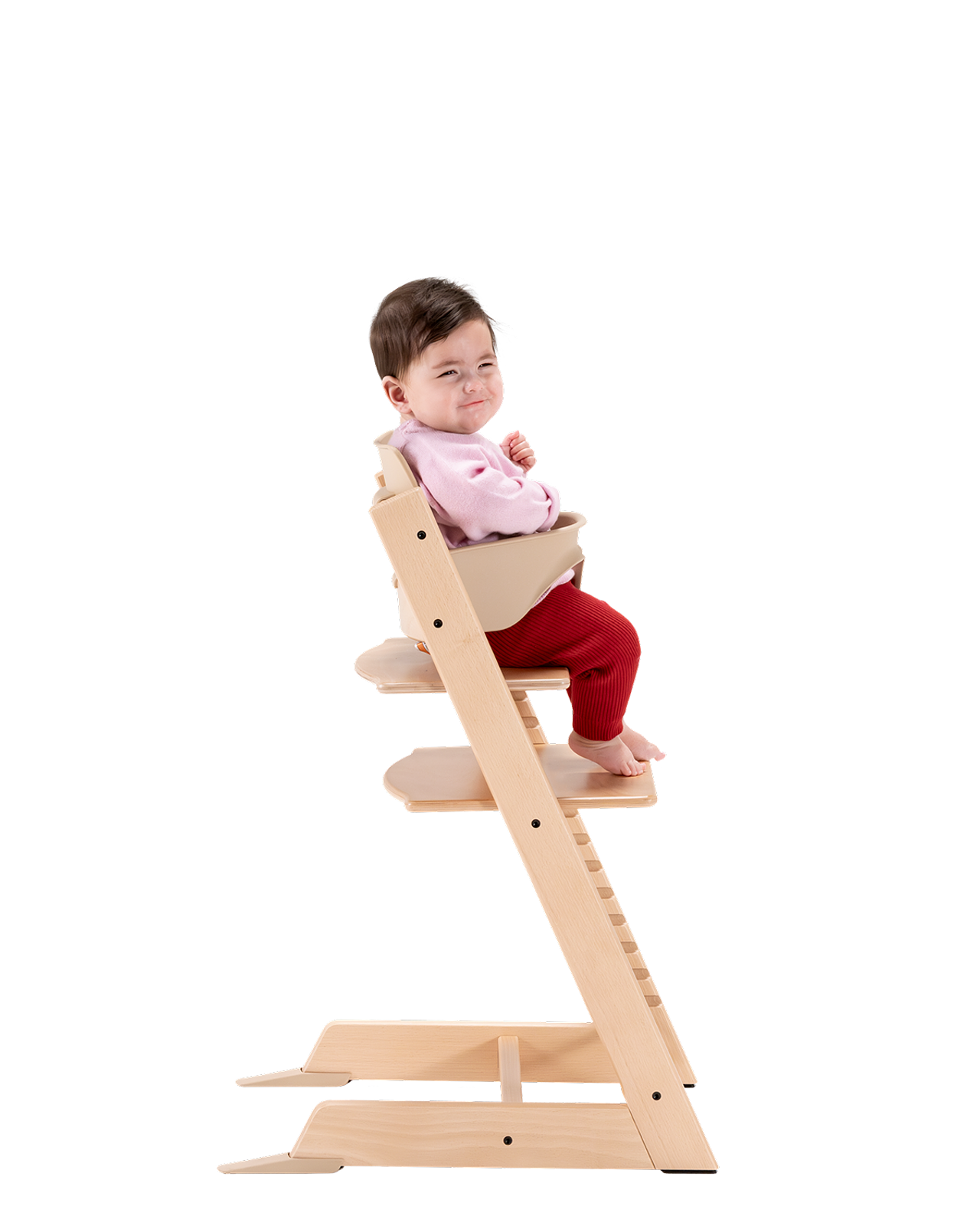 En bebis med ett snett leende sitter i en Tripp Trapp barnstol i naturträ med ett beige Baby Set.