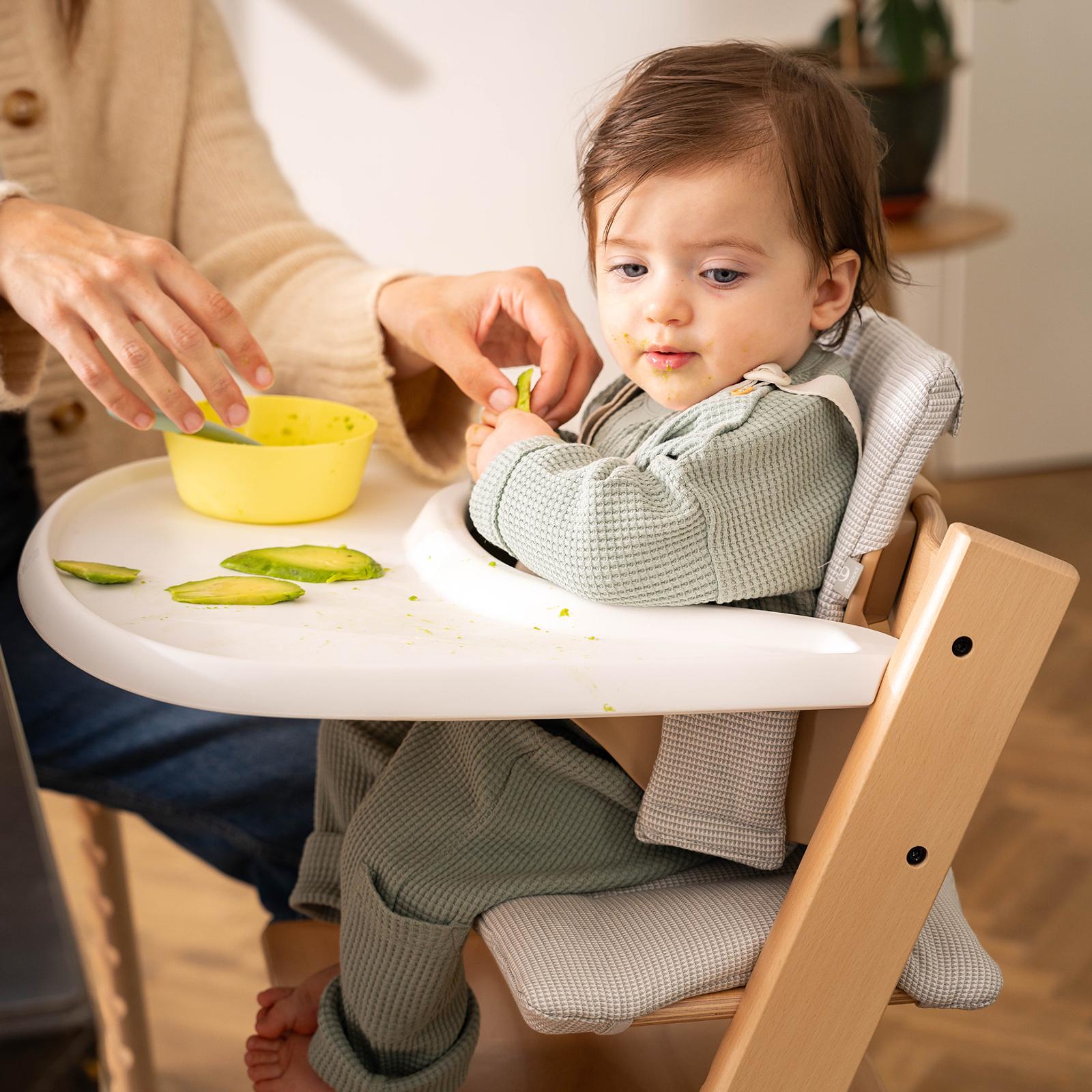 A baby with a messy face eats avocado in a Tripp Trapp high chair with an adult's help.