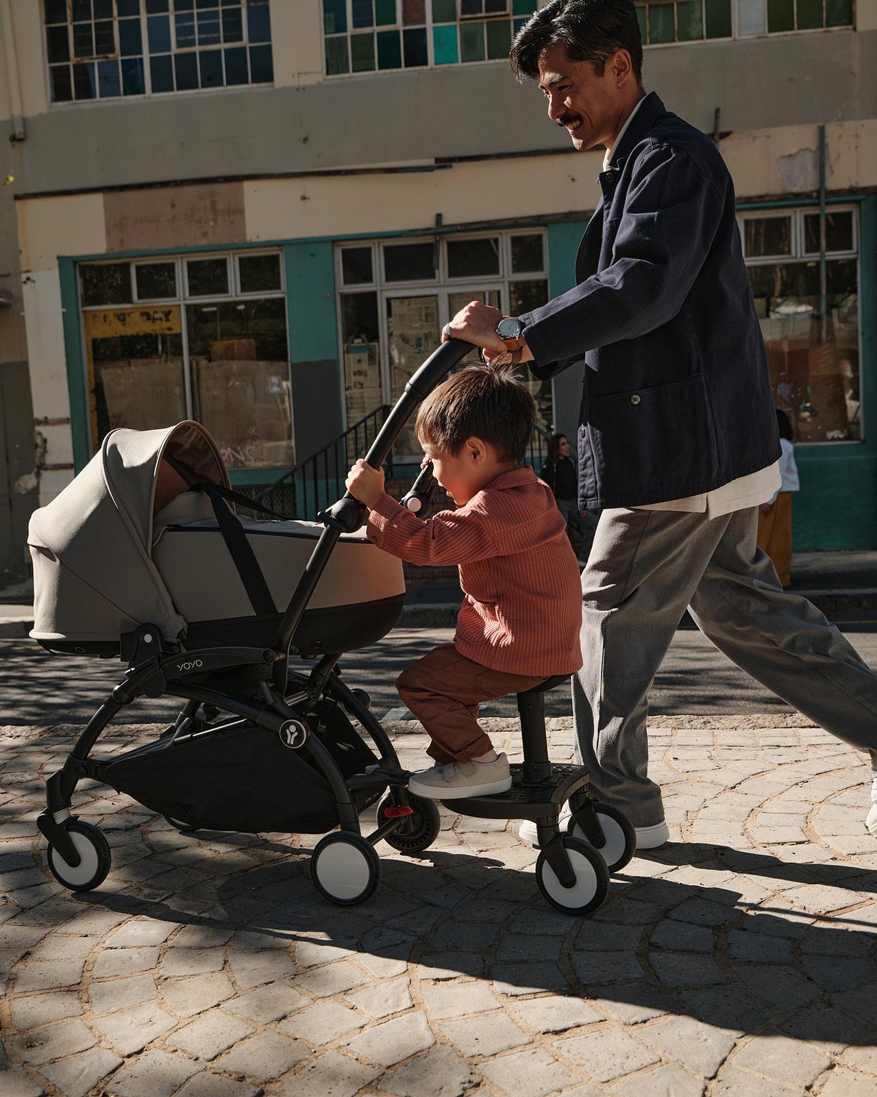 Un homme pousse une poussette STOKKE YOYO avec une nacelle et un enfant assis sur une planche à roulettes attachée.