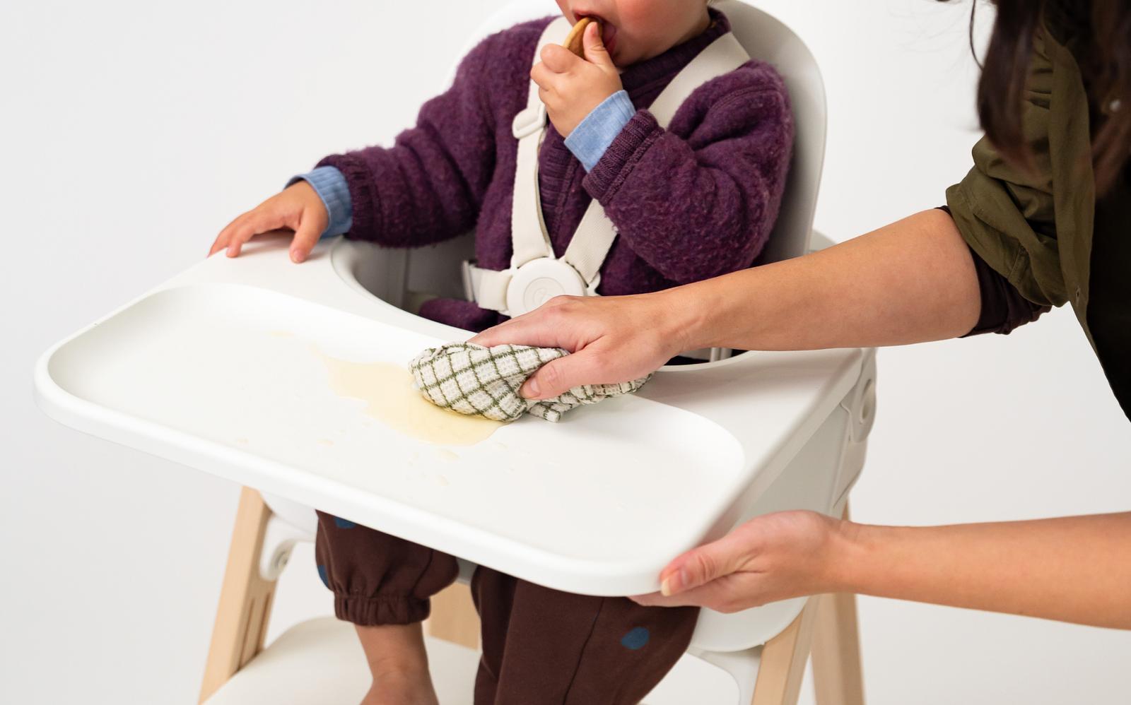 Child in Tripp Trapp high chair eating, while a hand cleans a spill on the tray.