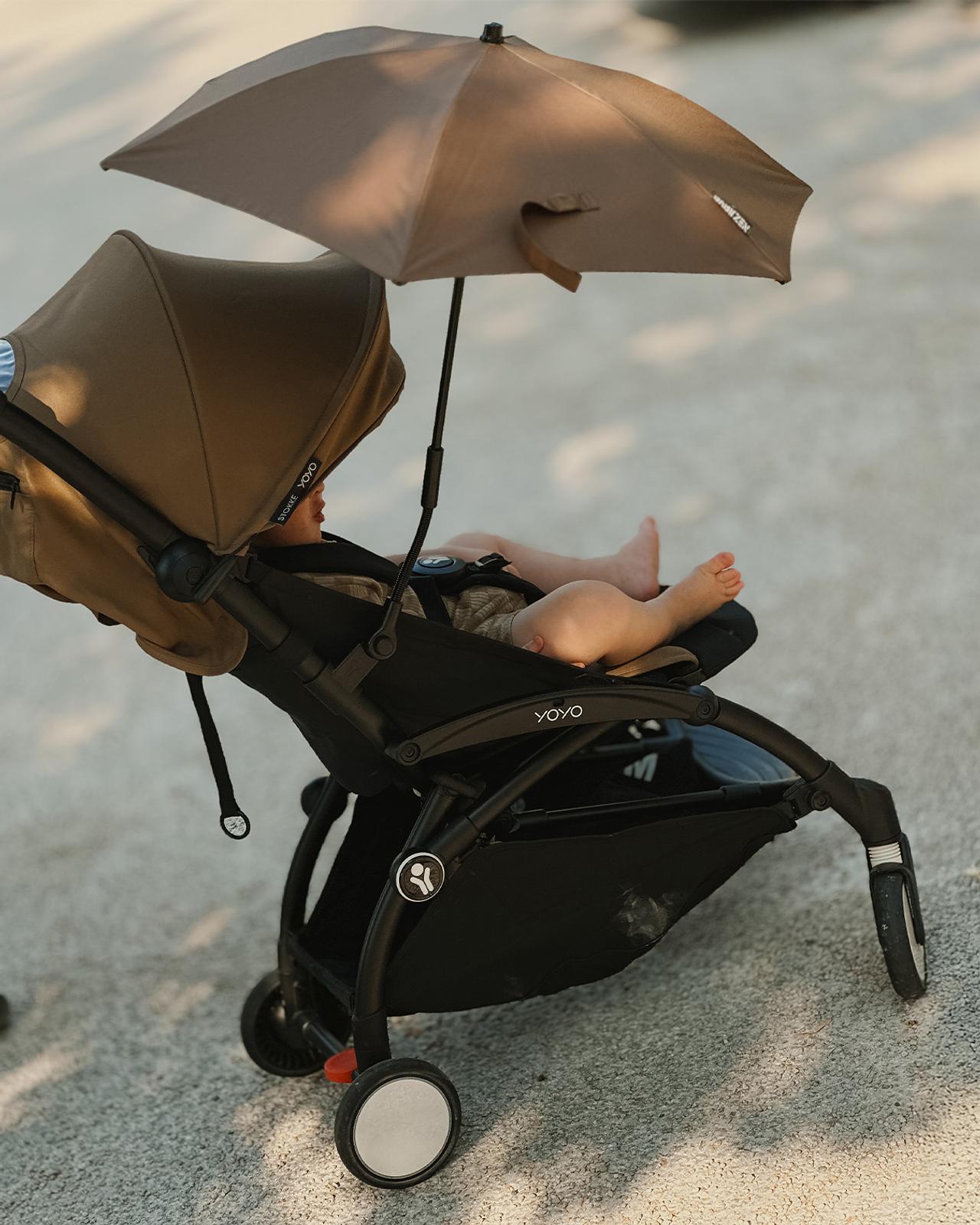 Baby in a brown STOKKE YOYO stroller with an attached brown umbrella, outdoors.
