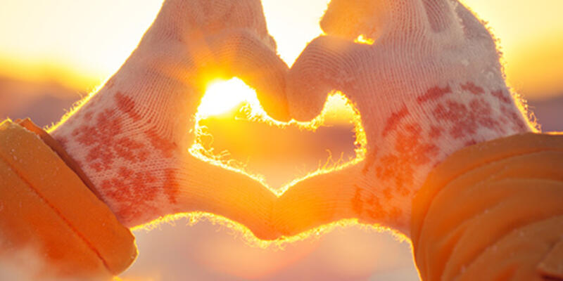 A woman wearing gloves outside making a heart with both hands while the sun is shining through.