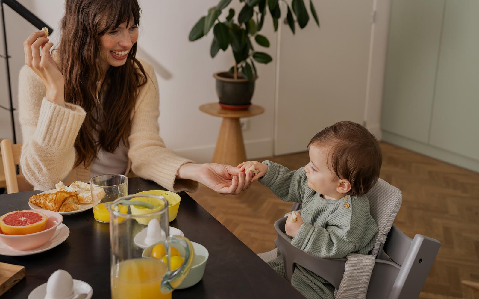 Leende kvinna erbjuder mat till bebis i en grå Tripp Trapp barnstol vid ett frukostbord.