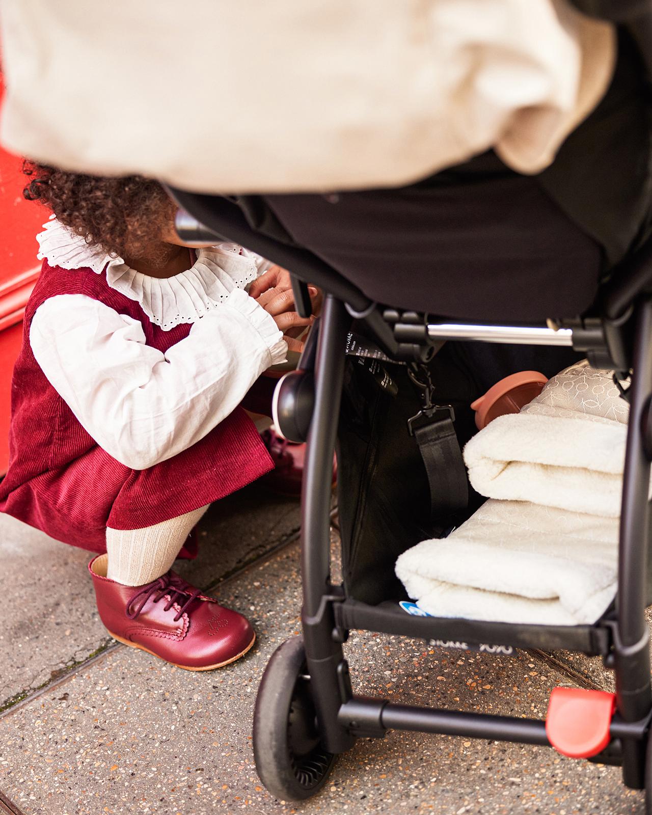 Enfant en robe rouge accroupi à côté d'une poussette STOKKE YOYO avec des couvertures dans son panier.