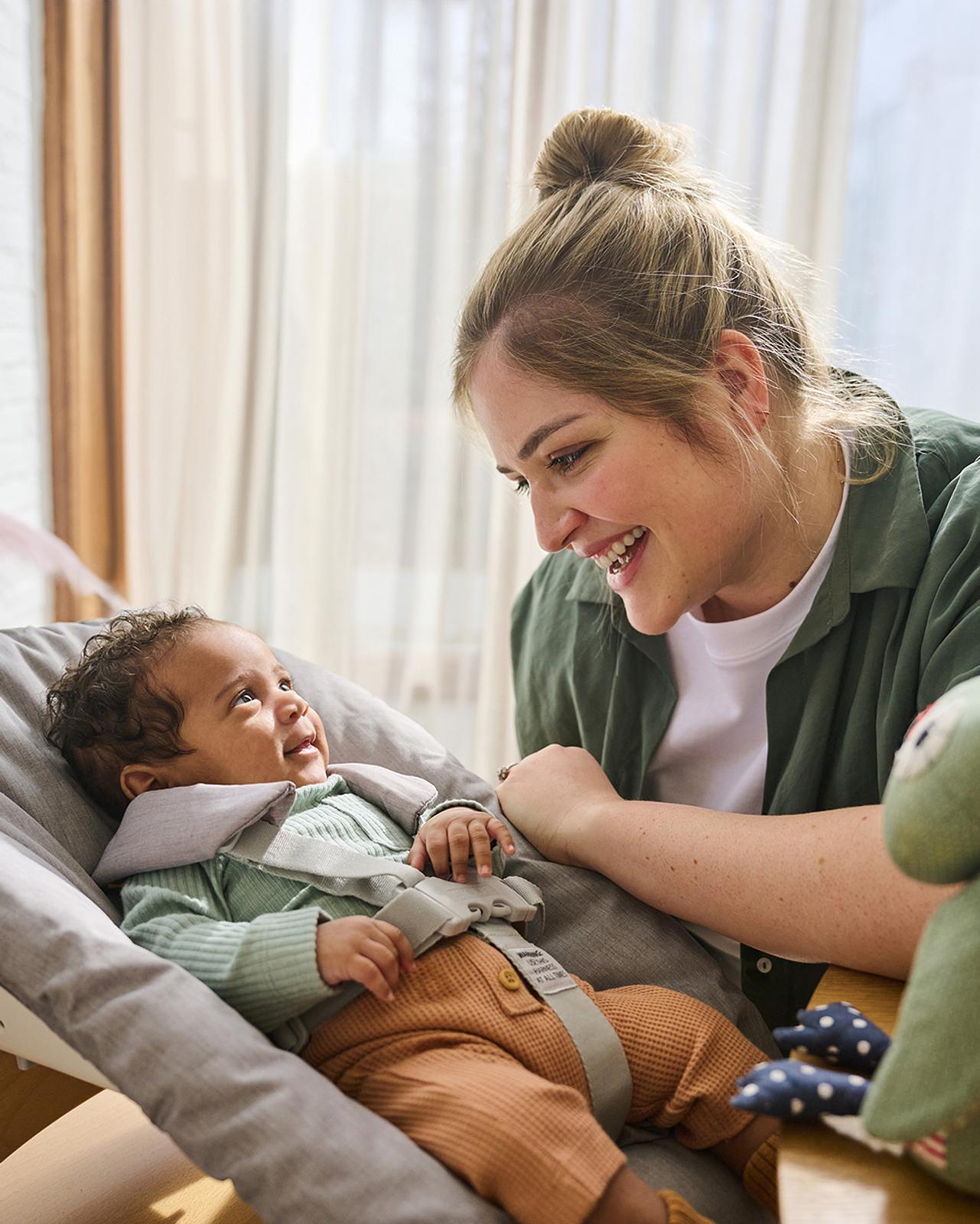 Una mujer sonriente mira con cariño a un bebé en una trona Tripp Trapp con un Newborn Set.