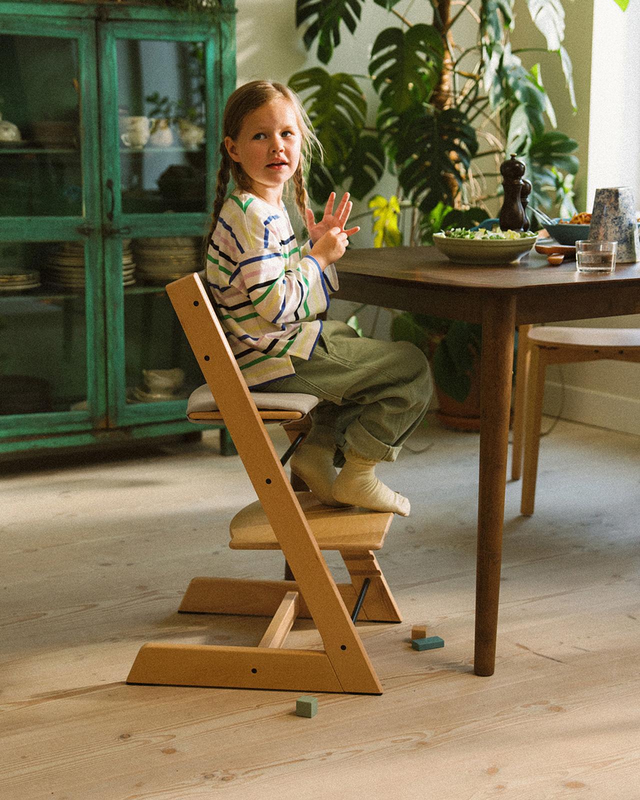 En ung flicka sitter vid ett träbord i en Tripp Trapp-stol i naturträ och pratar med händerna.