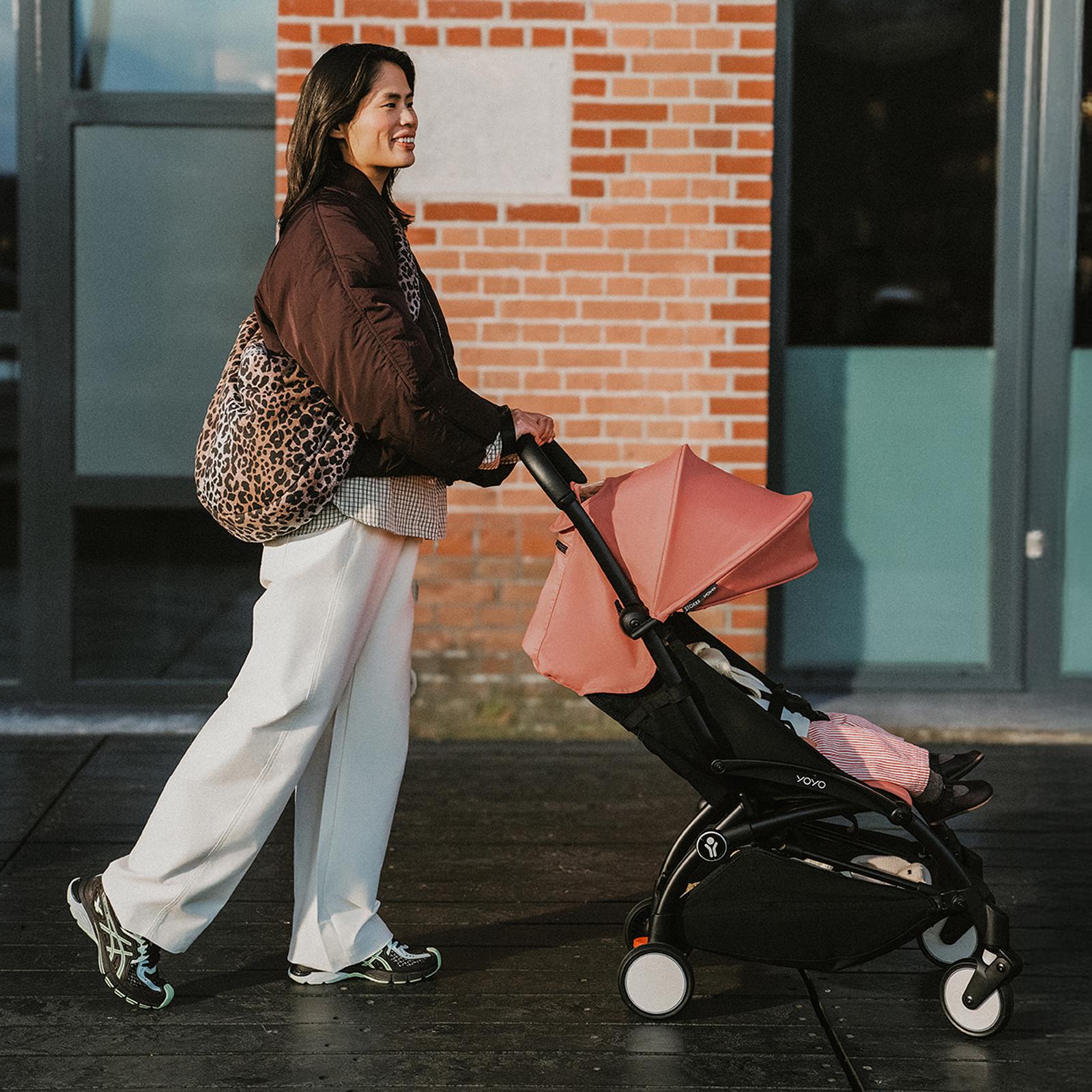 A smiling woman pushes a STOKKE YOYO stroller with a baby inside, outdoors on a sunny day.