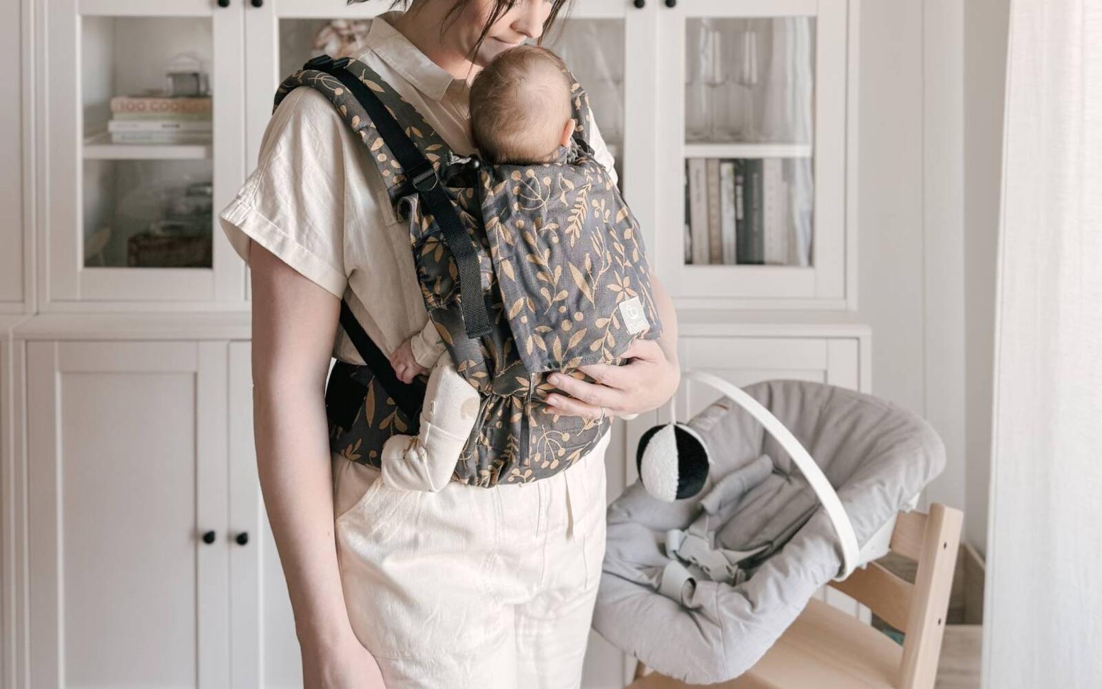 Mother using limas flex baby carrier in her home with the tripp trapp chair with newborn set is in the background.