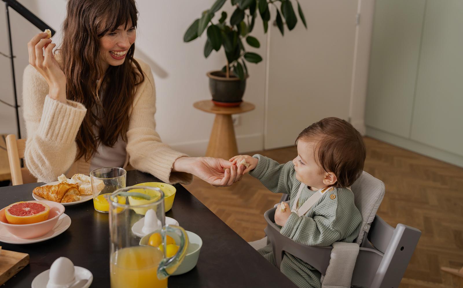 A smiling woman feeds a baby in a gray Tripp Trapp high chair at a breakfast table.
