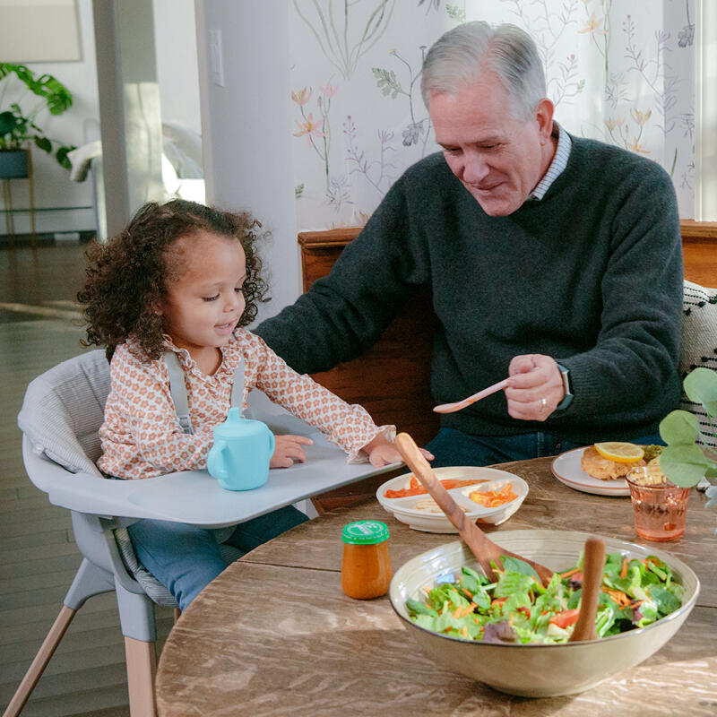Grandpa feeding his granddaughter at the kitchen table while she's sitting in her Clikk™ High Chair with tray and cushion.