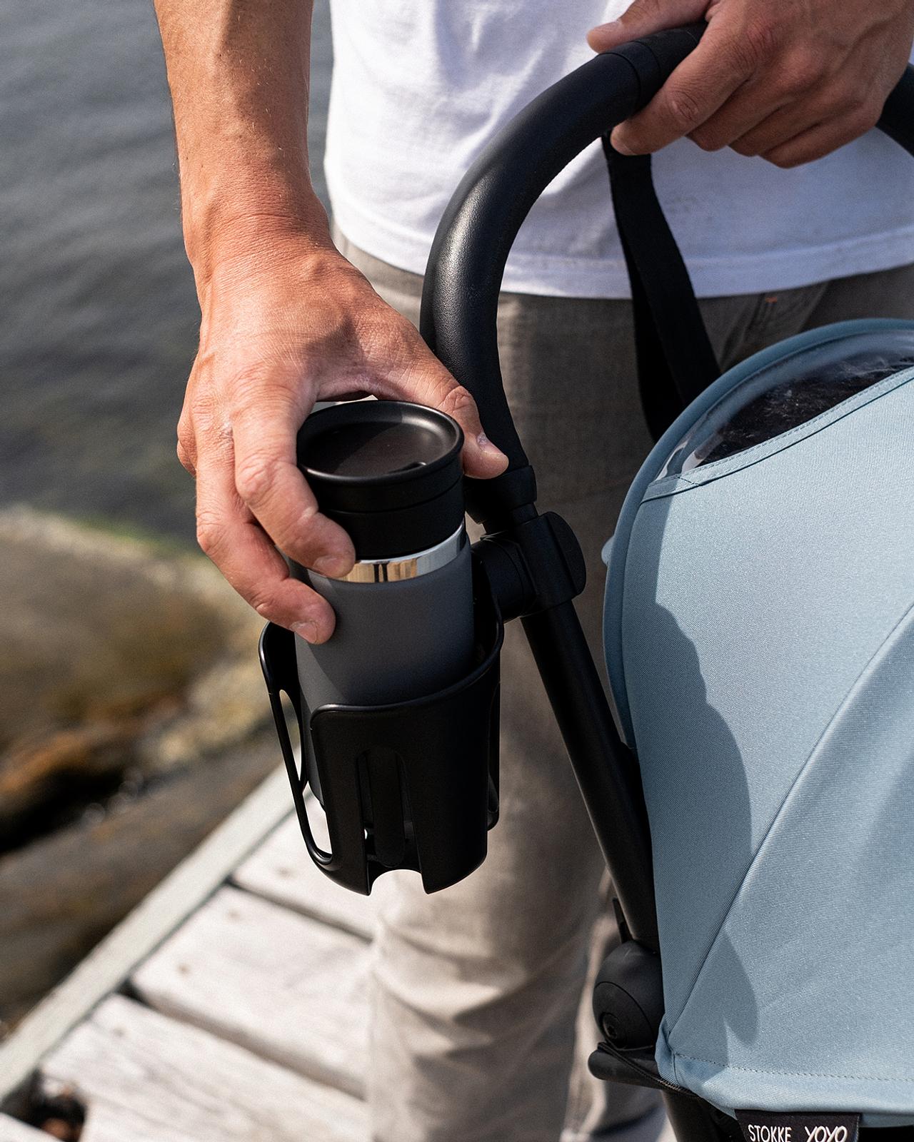 A hand holds a thermal cup in a STOKKE YOYO stroller's cup holder by the water.
