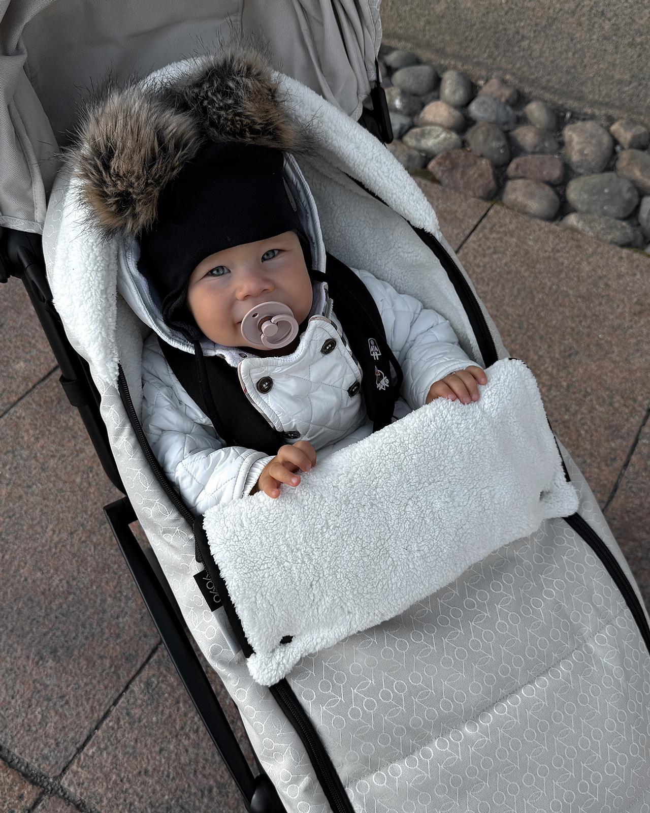 Un bébé coiffé d'un bonnet bordé de fourrure noire et vêtu d'une veste blanche est assis dans une poussette STOKKE YOYO, avec une tétine.