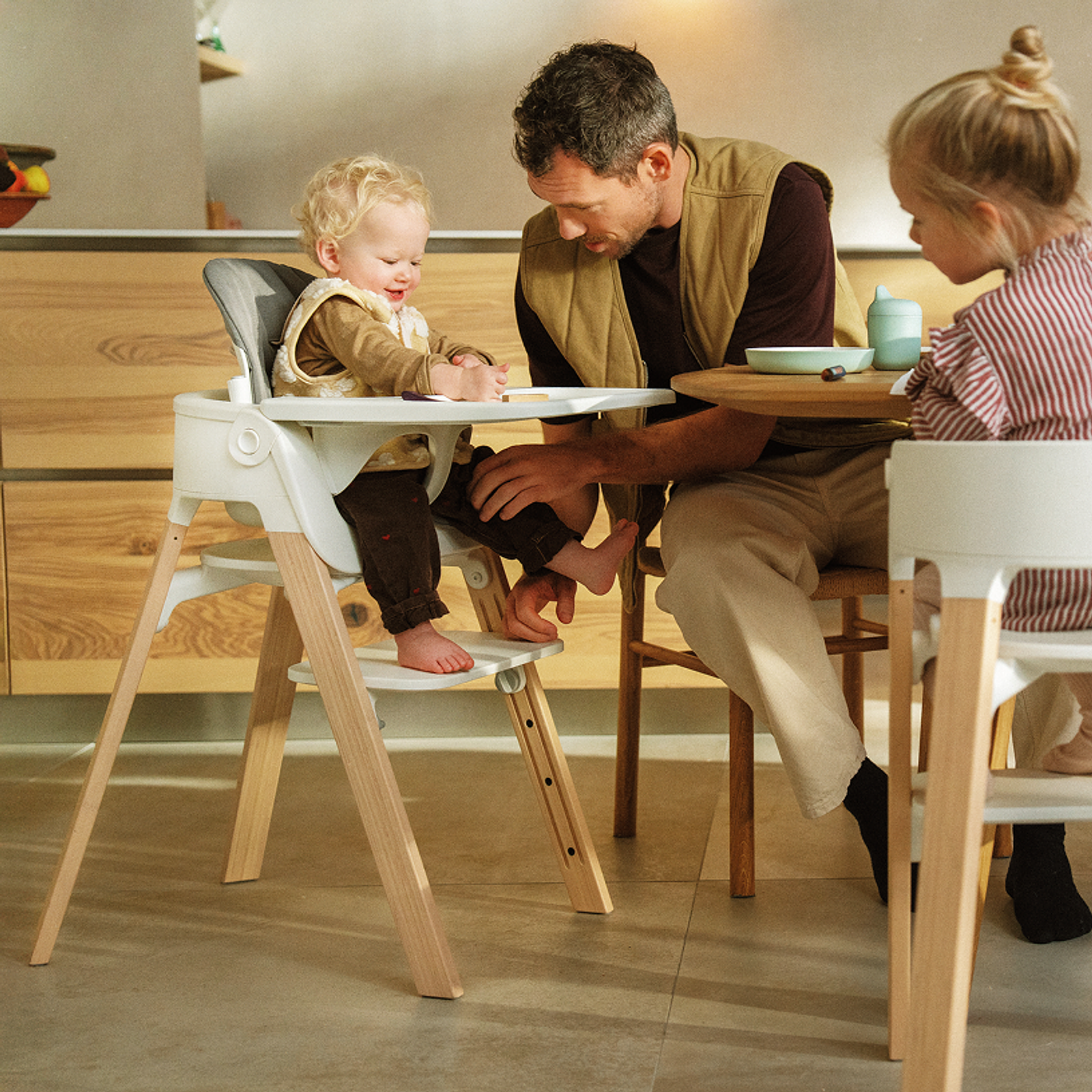 Father and two children at a wooden table. Baby smiles in a white high chair with wooden legs.