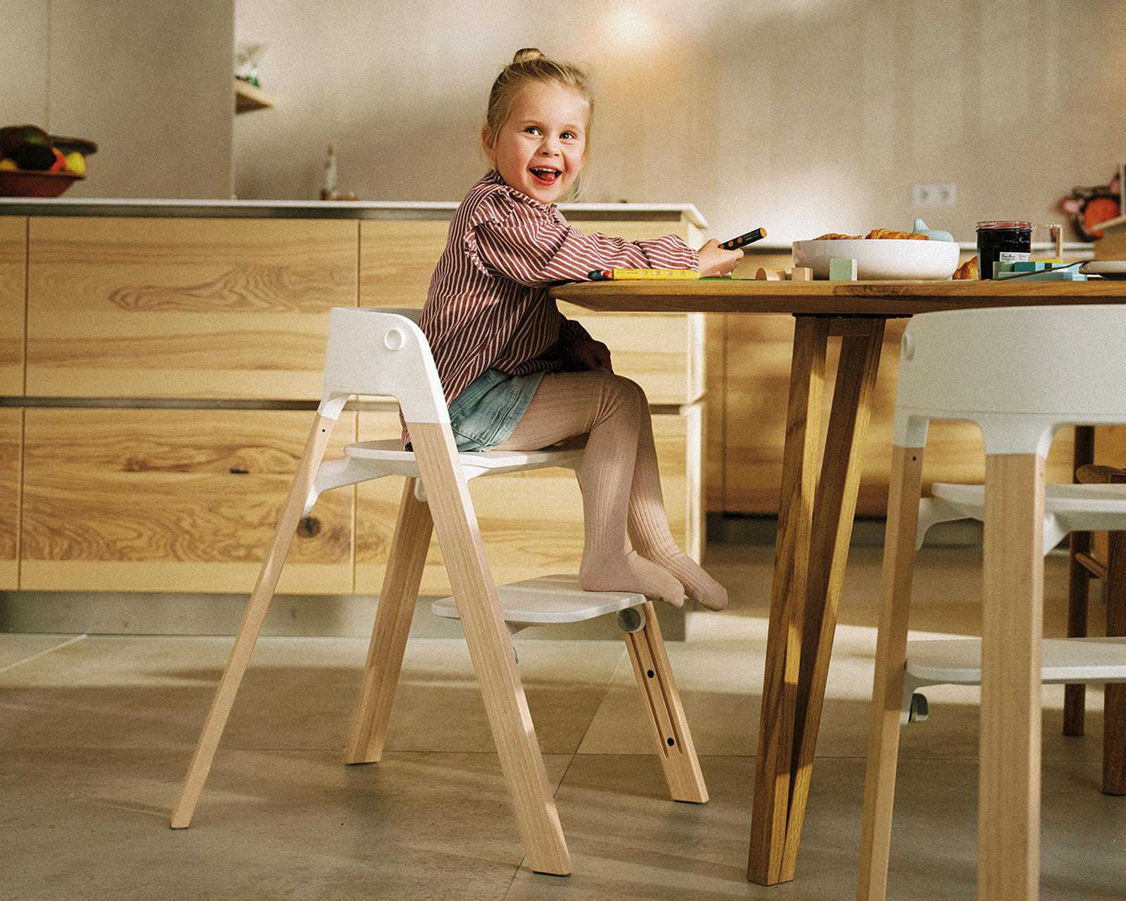 A smiling child sits at a wooden table in a white and wood high chair.