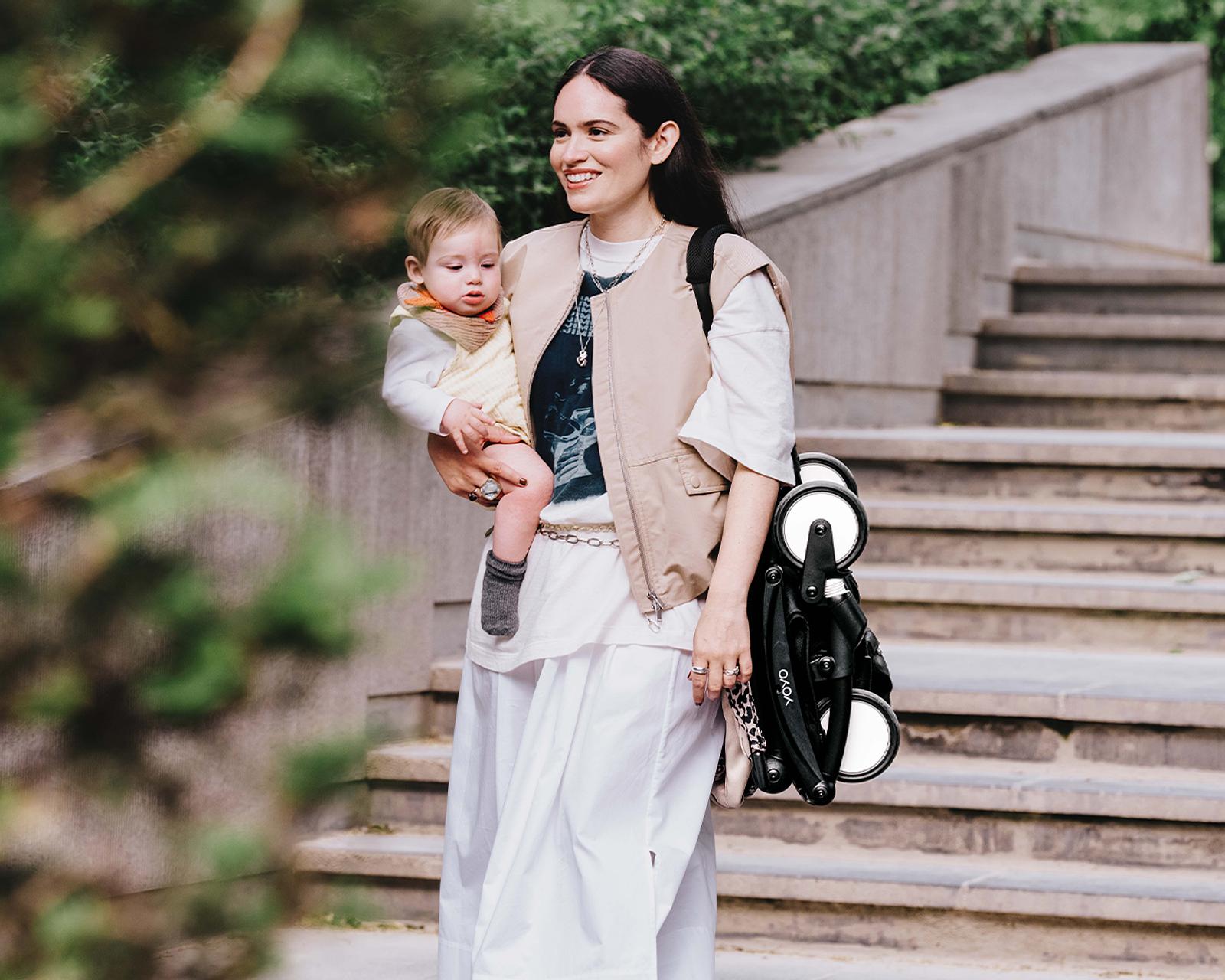Woman holds a baby and a folded STOKKE YOYO stroller, smiling outdoors near stairs.