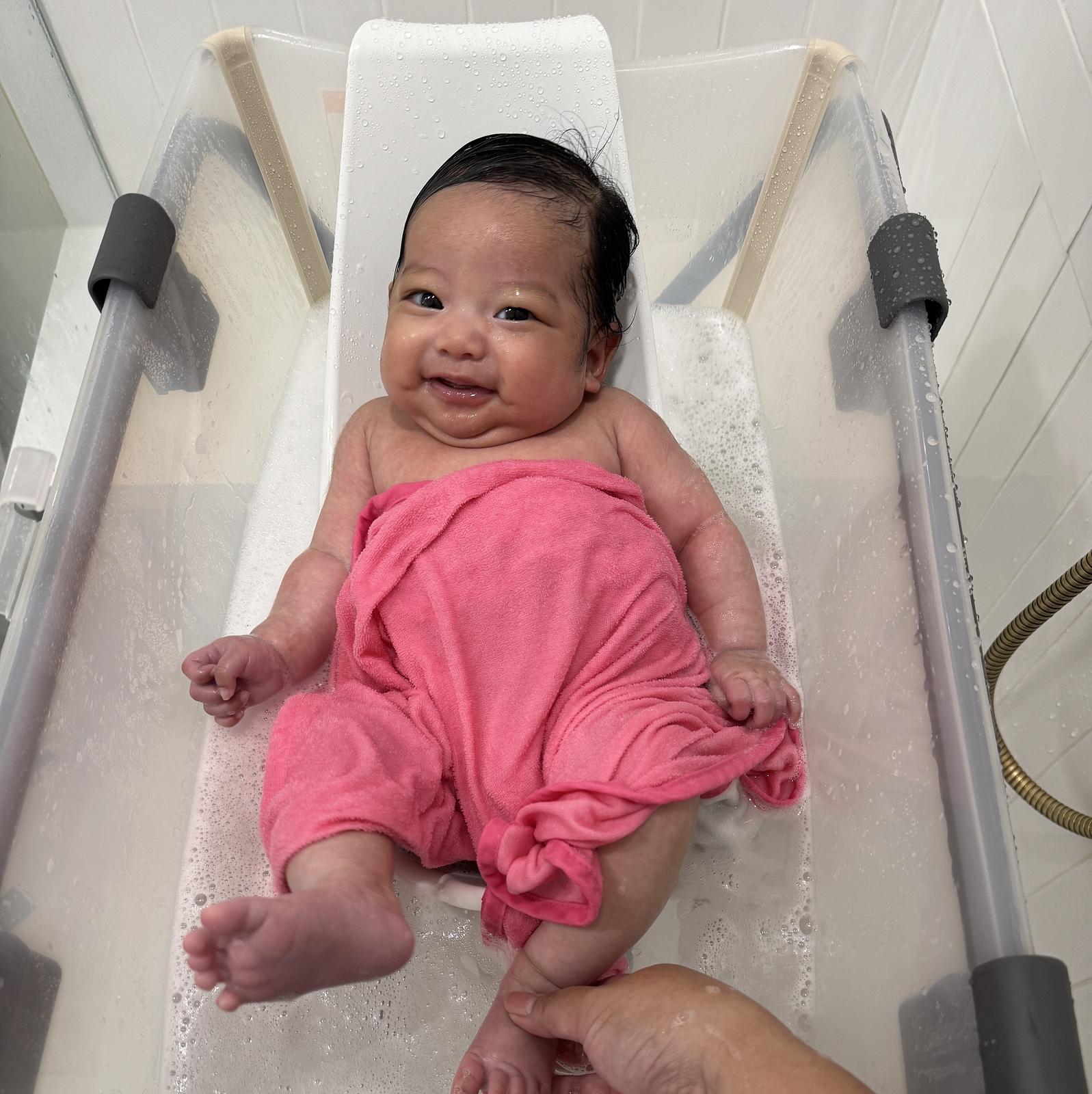 Smiling baby covered in a pink towel, being bathed in a STOKKE Flexi Bath with newborn support.