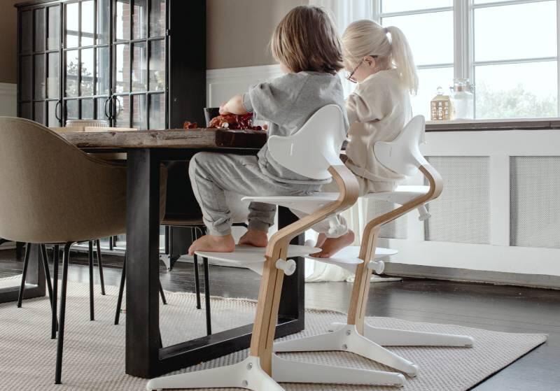 Two children sitting at the dining table on Stokke® Nomi® chairs.