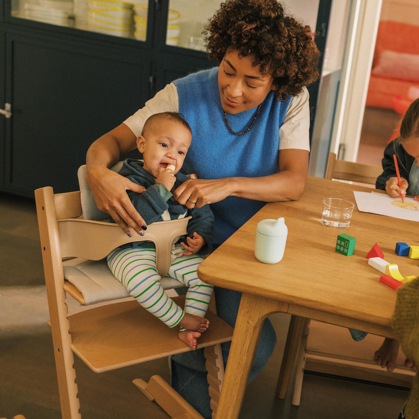 A woman helps a baby eating in a Tripp Trapp high chair at a wooden table.