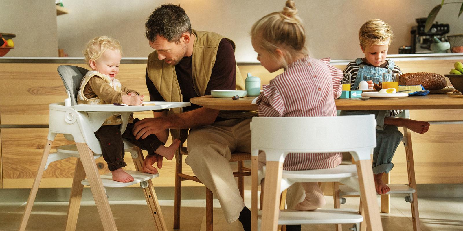 Un père aide un bébé souriant assis dans une chaise haute blanche à une table avec deux autres enfants.