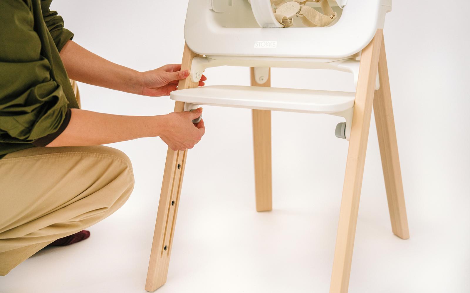 Hands adjust the seat of a white and wooden high chair.