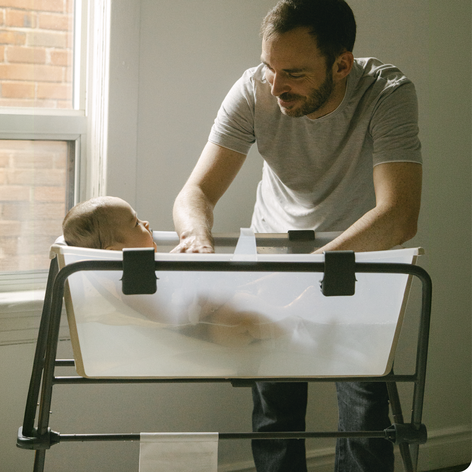 A father gently bathes his baby in a transparent baby bath on a stand near a window.